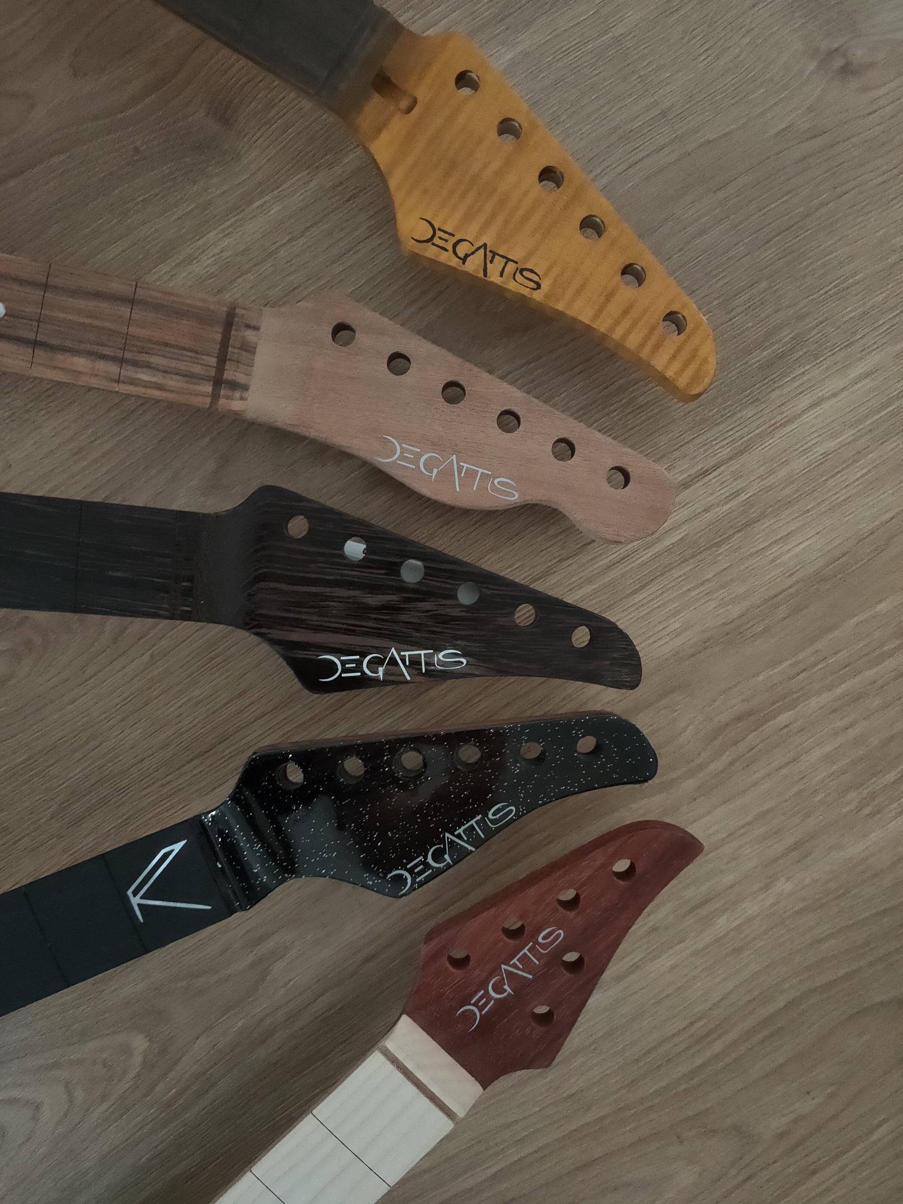 Five guitar headstocks in different woods, all carrying the De Gattis mark